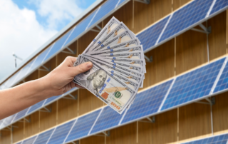A person's hand holding Money fanned out in front of solar panels.
